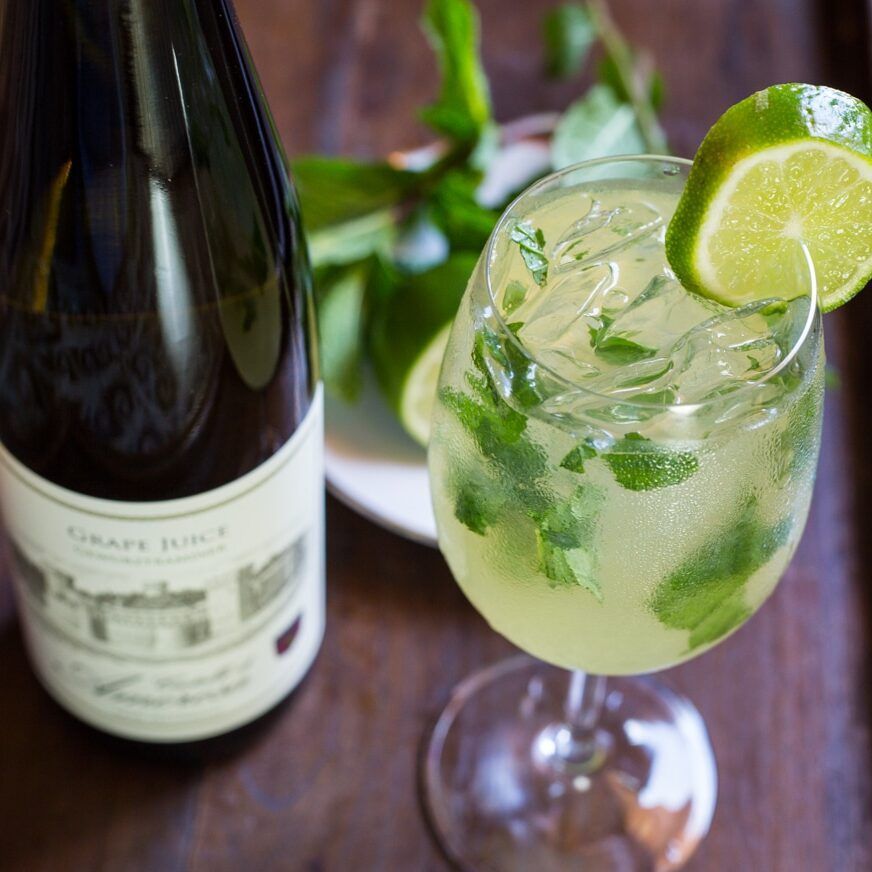 Cocktail with lime and mint, next to a grape juice bottle on a wooden tray.
