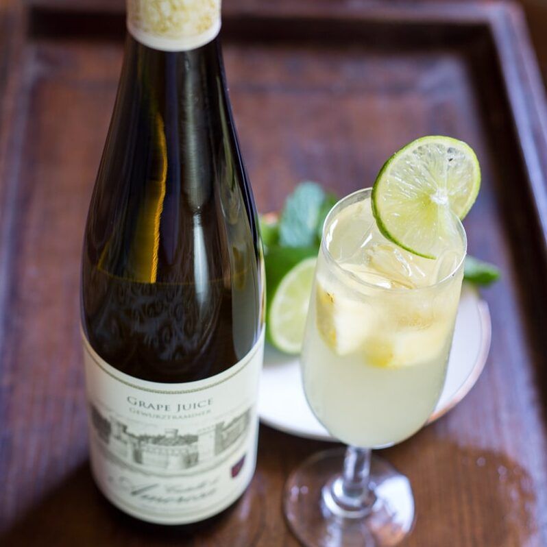 Bottle of grape juice next to a glass with ice and lime garnish on a wooden tray.