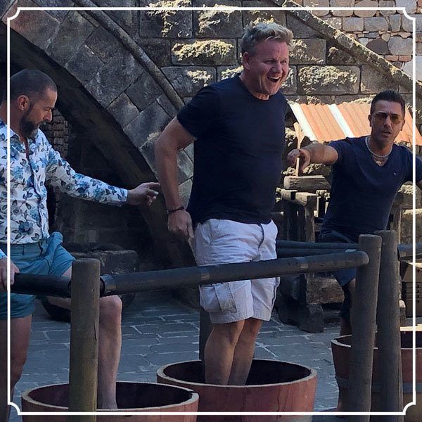 Three men stomping grapes in wooden vats. Gordon Ramsay laughs, arms raised. Outdoors, stone backdrop.