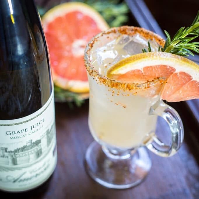 Cocktail with grapefruit garnish, spicy rim, beside a bottle of grape juice and a halved grapefruit.