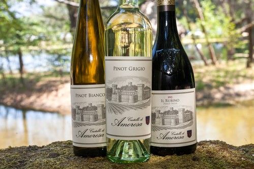 Three Castello di Amorosa wine bottles displayed outdoors near water and trees