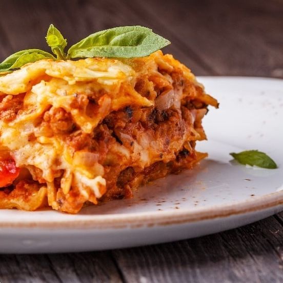 Lasagna slice on a white plate, topped with basil, on a wood surface.