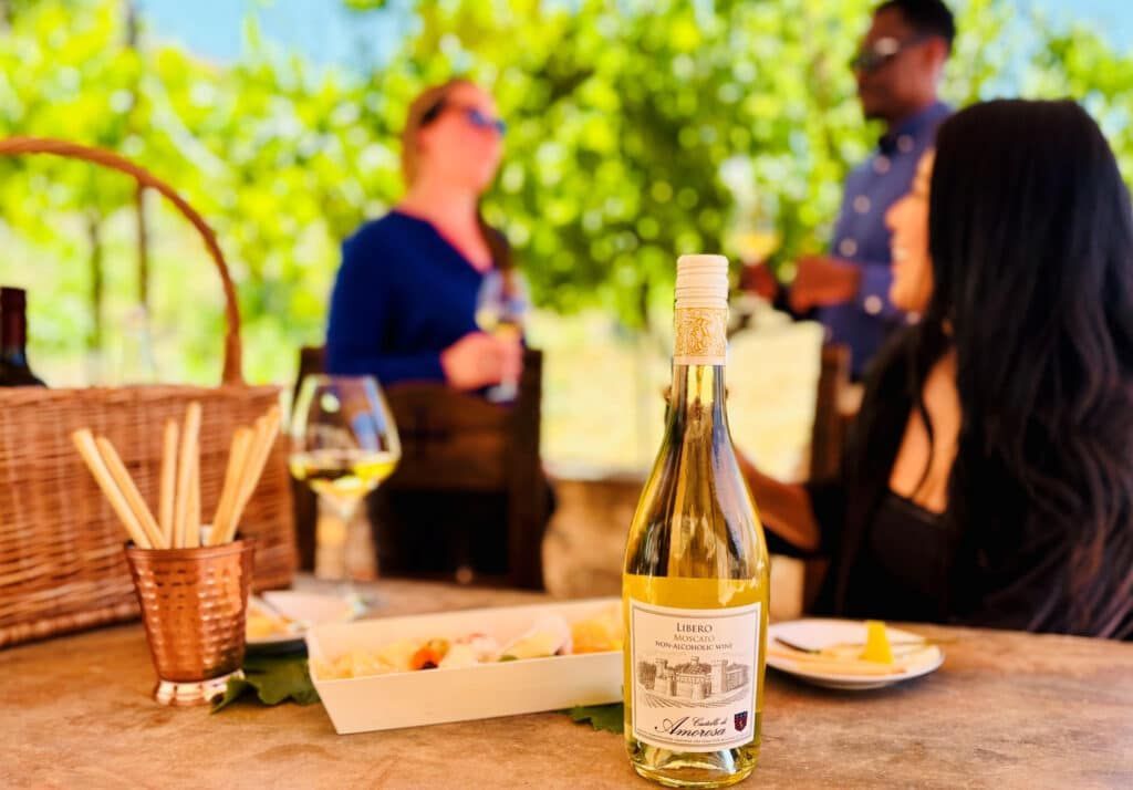 Wine bottle and snacks on a table at a vineyard with people enjoying drinks in the background.