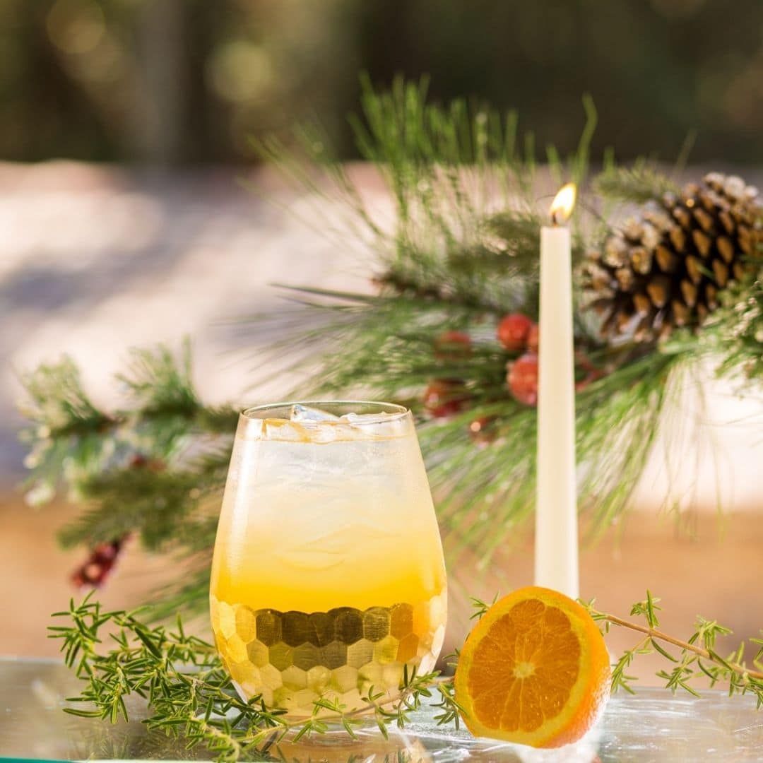 Cocktail in textured glass, orange slice, candle, and pine bough on table.