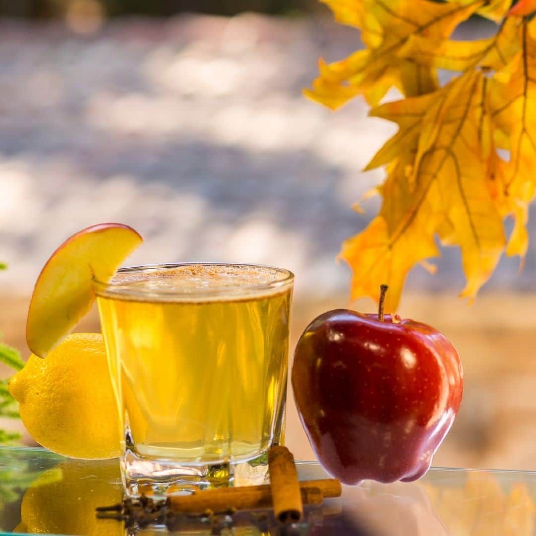 Glass of apple cider with apple slice garnish, red apple, lemon, cinnamon, fall leaves.