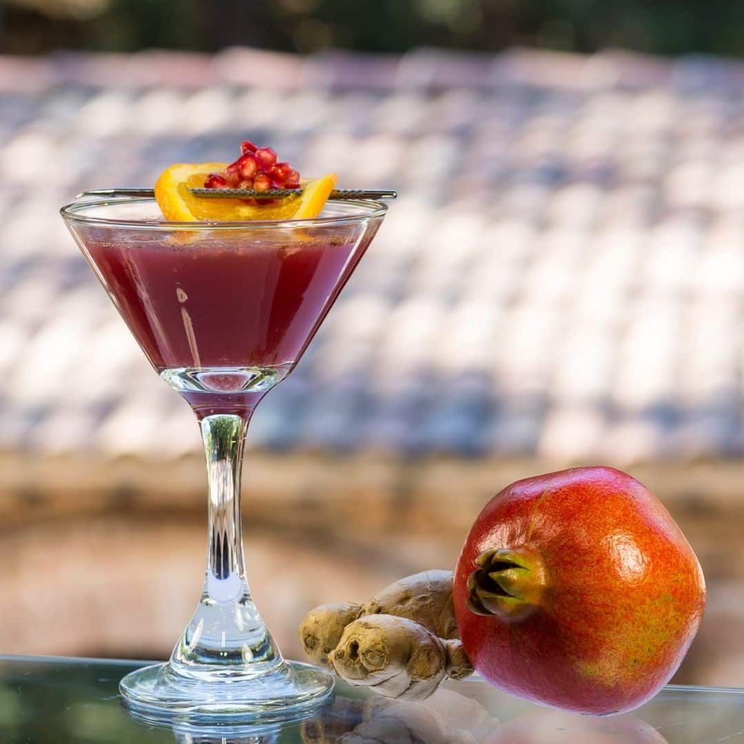 A red cocktail in a martini glass garnished with pomegranate seeds and orange peel, alongside a pomegranate and ginger.