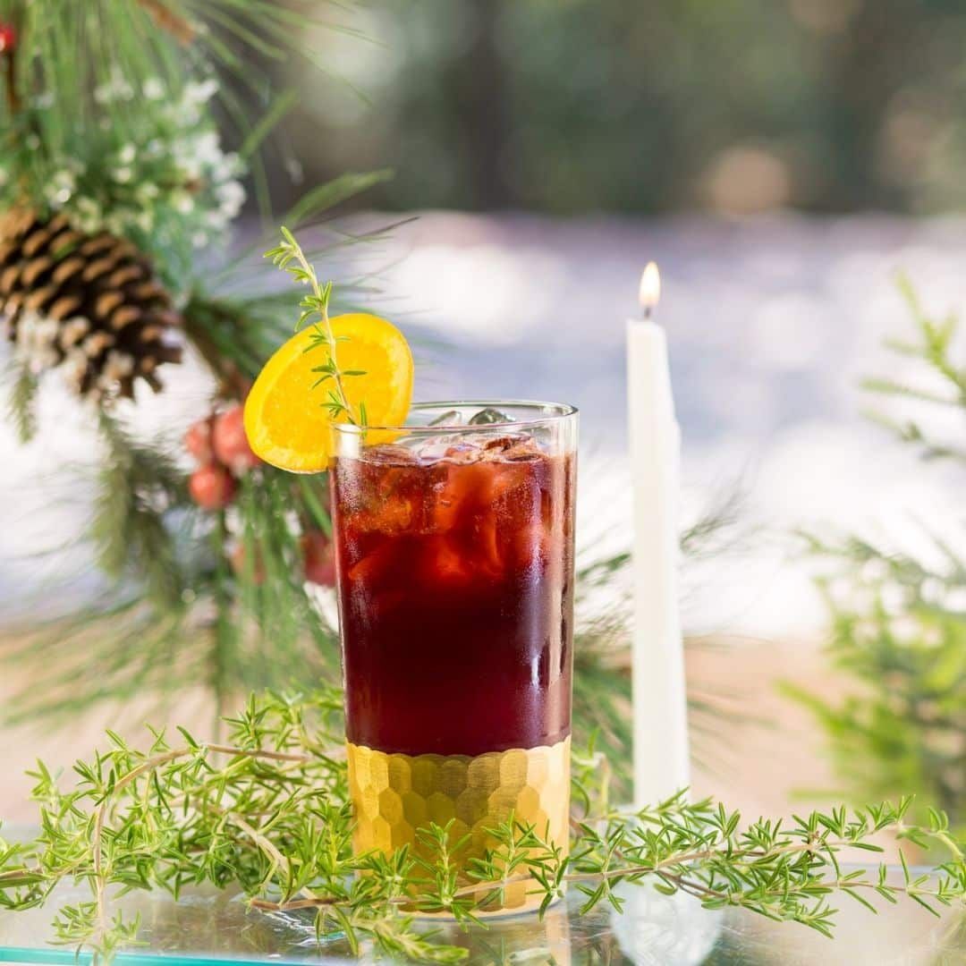 Cocktail in a golden-bottomed glass, garnished with orange and thyme, on a table near greenery and a candle.