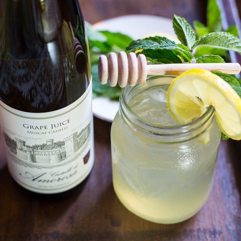 Glass of grape juice with lemon and mint, beside a bottle and honey dipper.