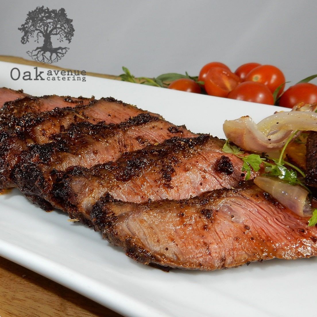 Sliced grilled steak on a white plate with tomatoes and onions; catering logo.
