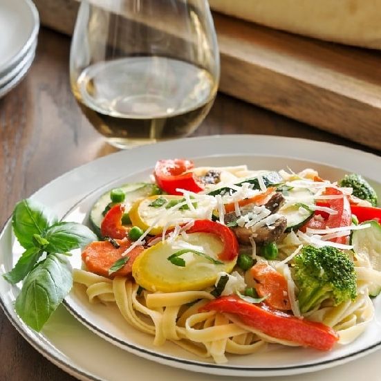 Pasta with vegetables, including zucchini, carrots, and tomatoes, topped with cheese, next to a glass of white wine.