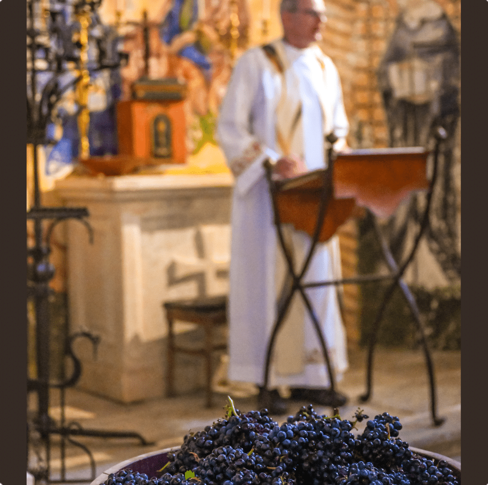 Bowl of dark grapes in foreground, priest in white robe at lectern in church.