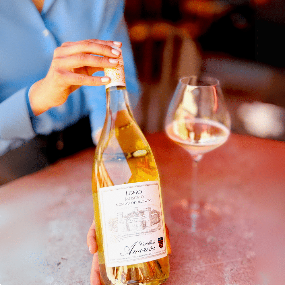 Person opening a bottle of white wine with a full glass next to it on a table.