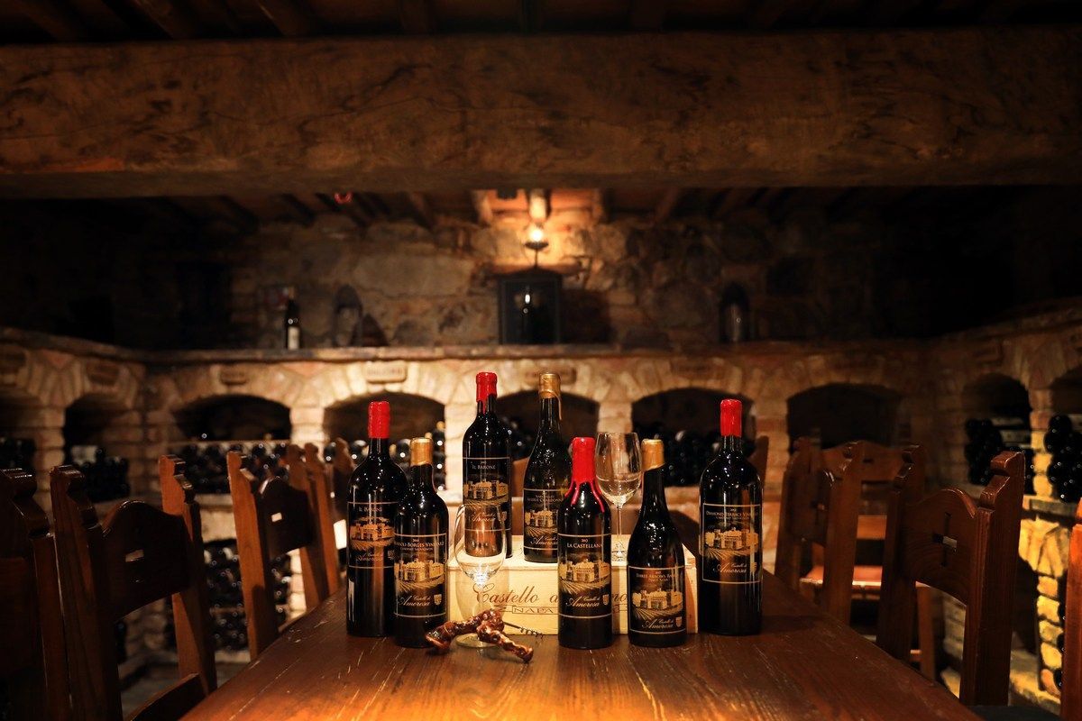 Wine bottles on a wooden table in a dimly lit wine cellar with stone walls and wooden chairs