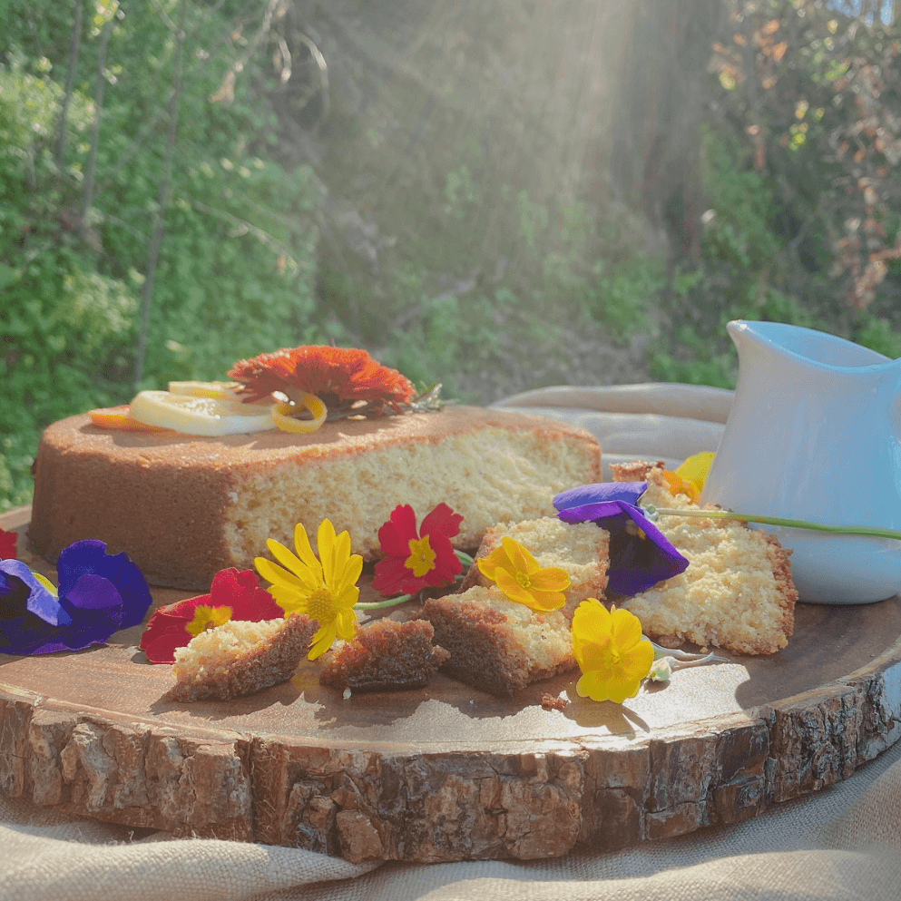 Cake decorated with flowers on a wooden platter, outdoors with a pitcher of liquid.