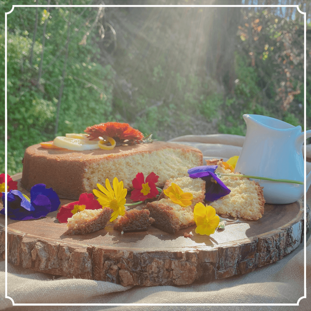 Cake decorated with flowers on a wooden platter outdoors, with pitcher and sunlit background.