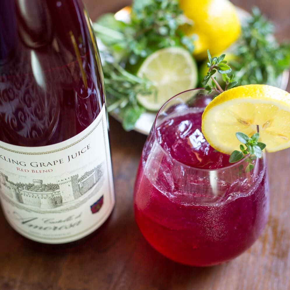 Bottle of sparkling grape juice next to a glass filled with a purple drink, garnished with lemon and herbs.