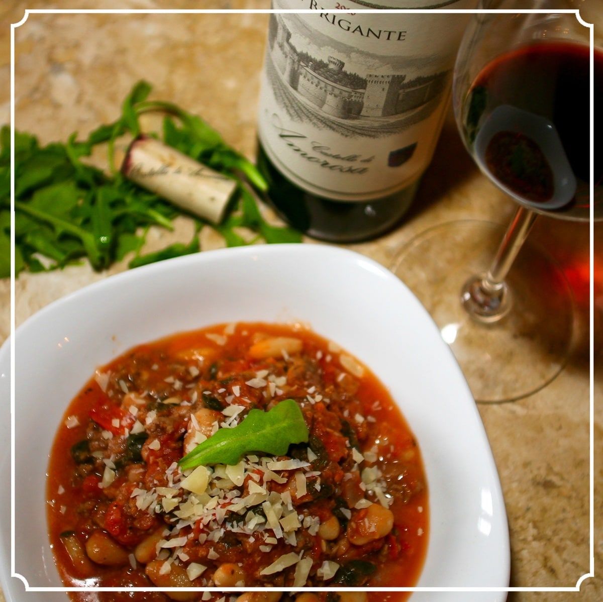 Soup in white bowl with wine glass, bottle, and arugula.