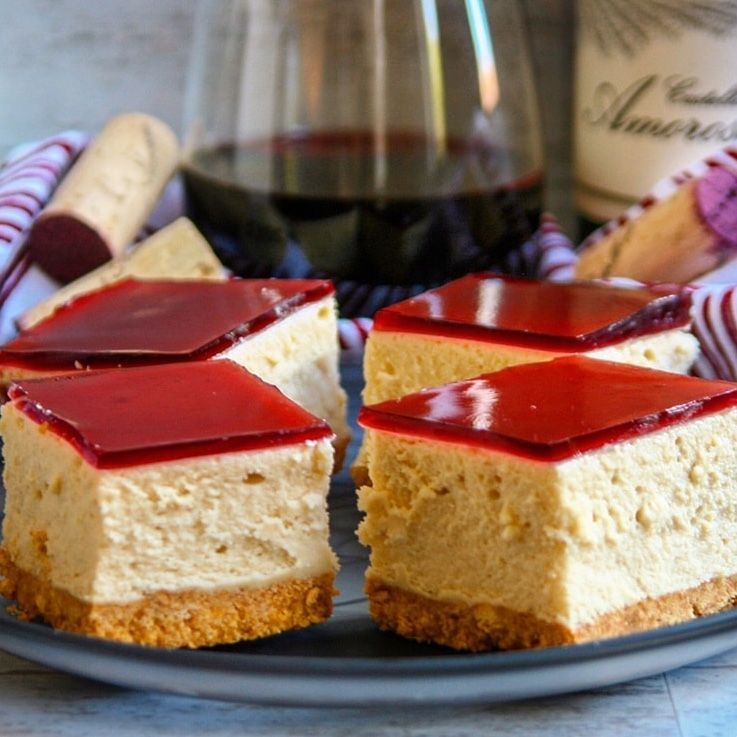 Cheesecake squares on a plate with red topping and wine in the background.