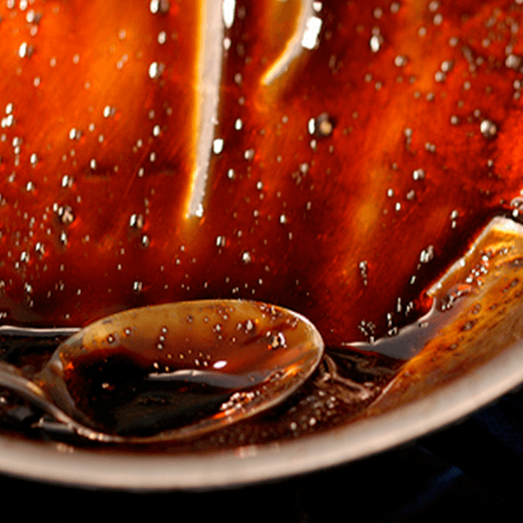 Spoon in dark, glossy sauce, possibly caramel or glaze, inside a white bowl.