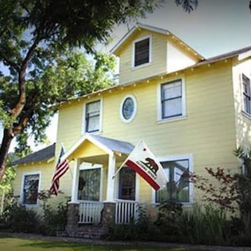 Yellow two-story house with American and California flags out front, surrounded by trees and greenery.