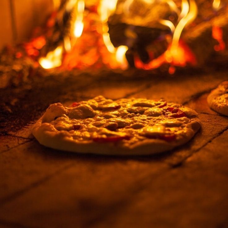 Pizza baking in a wood-fired oven with flames in the background.