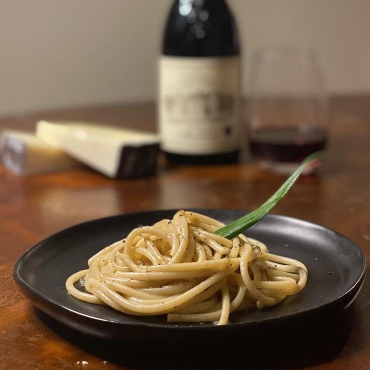 Pasta with pepper, served on a black plate, next to cheese and a bottle of red wine.