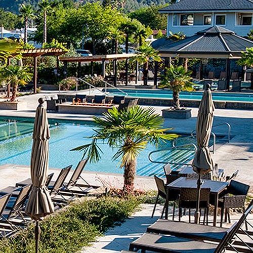 Poolside scene with lounge chairs, tables, palm trees, gazebo, and multi-story building in the background.