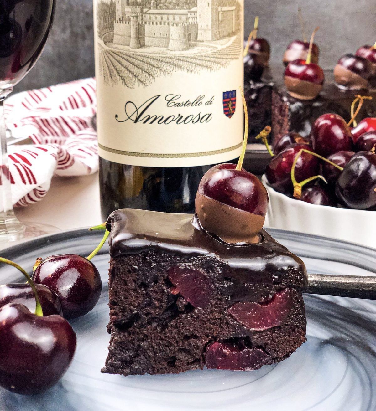 Slice of chocolate cake with cherries, bottle of Castello di Amorosa wine, fresh cherries, wine glass.