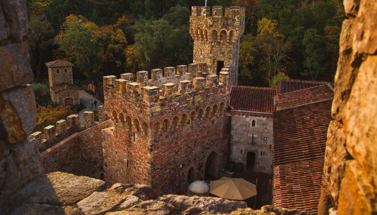 Stone castle with crenellated walls, a round tower, and a red tile roof, set on a rocky landscape.