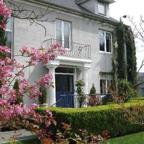 Gray house with blue door, balcony, and flowering tree in the foreground. Green hedge and shrubbery.