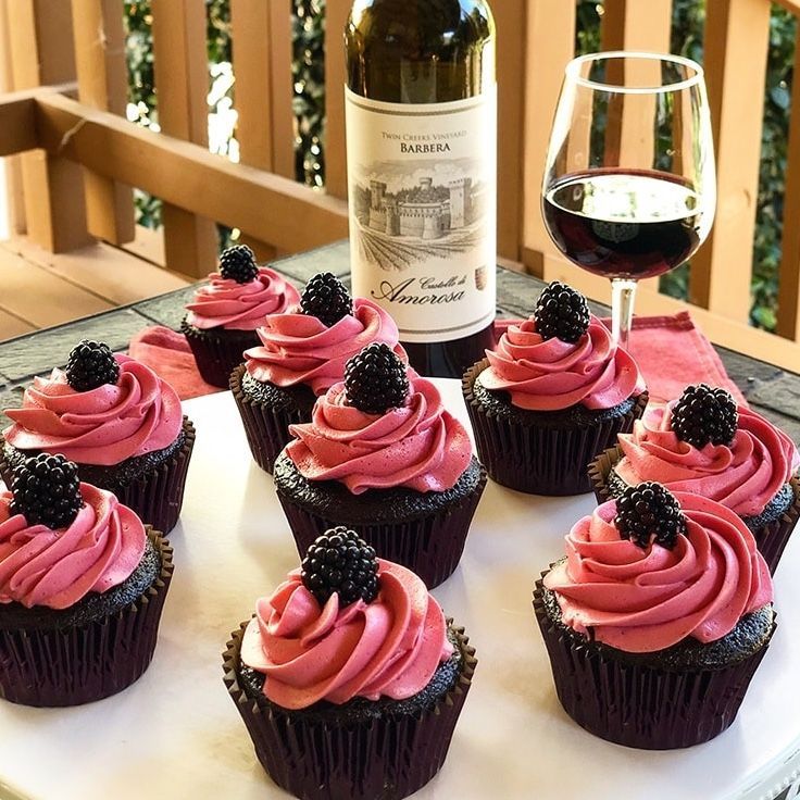 Dark cupcakes with pink frosting topped with blackberries, wine, and a bottle on a patio.