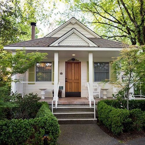 White cottage with brown door, porch, and steps, surrounded by green shrubs and trees.