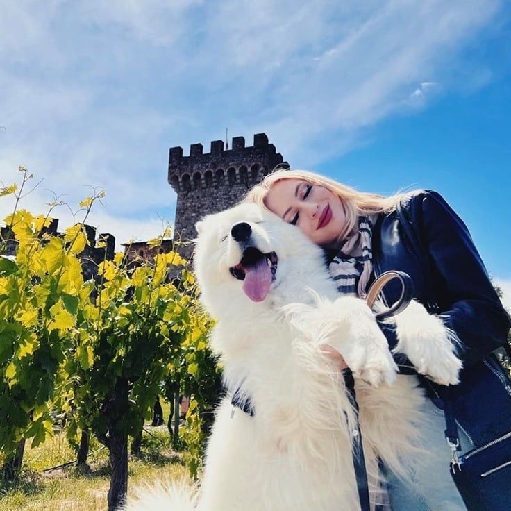 Woman hugging a large white dog in a vineyard with a stone tower in the background, blue sky.