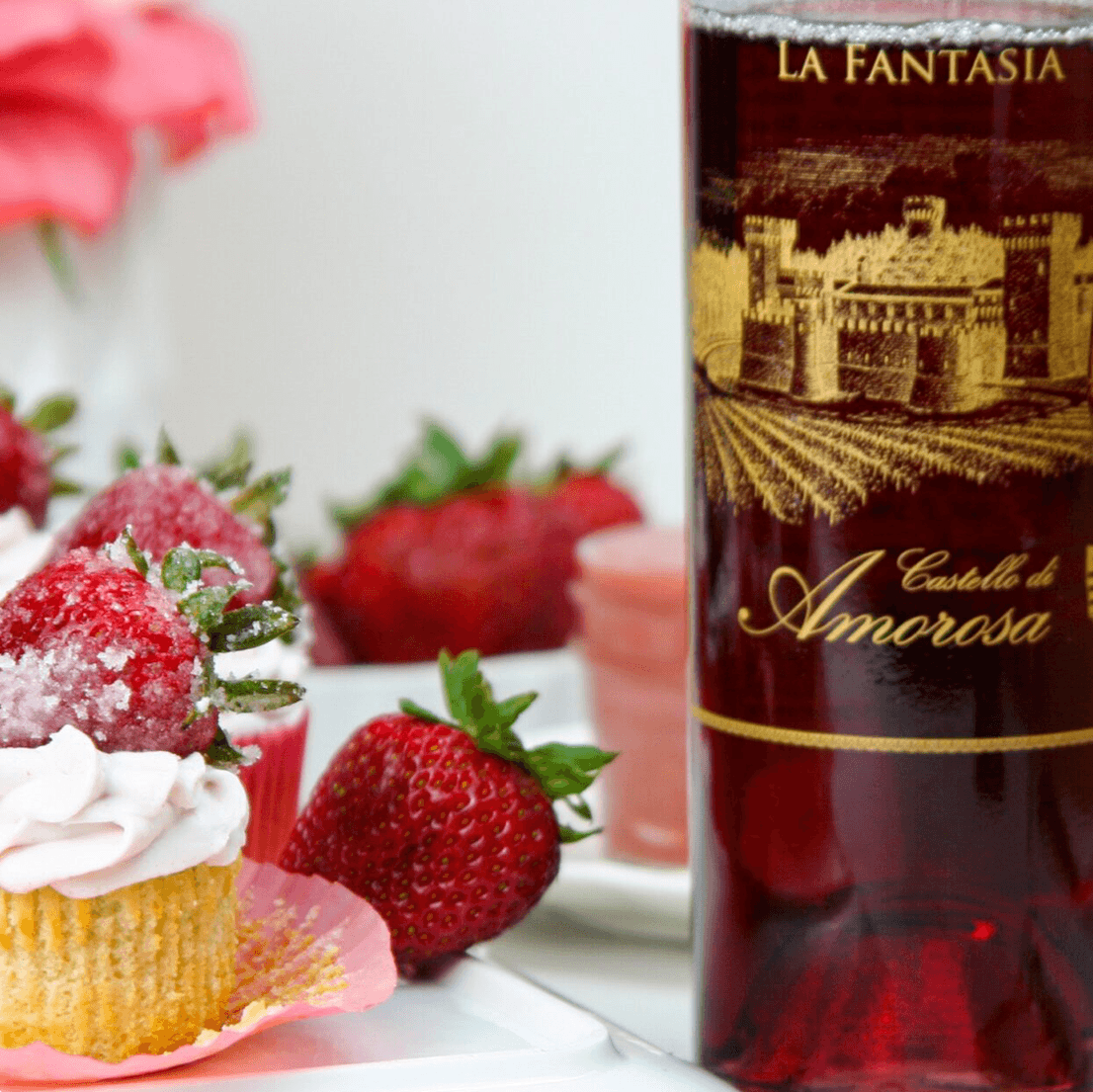 Bottle of red wine, cupcakes, and strawberries, arranged on a table.