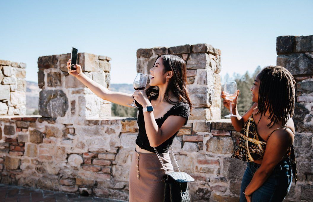 Two women taking a selfie on a stone wall, one holding a glass of red wine, outdoors in sunny weather.
