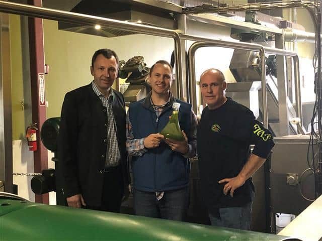 Three men in an olive oil production facility, one holding a green bottle.