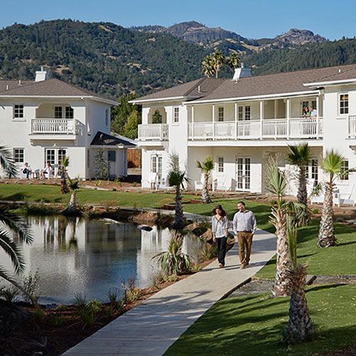 People walking near a pond in front of white buildings with mountain backdrop. Palm trees are present.