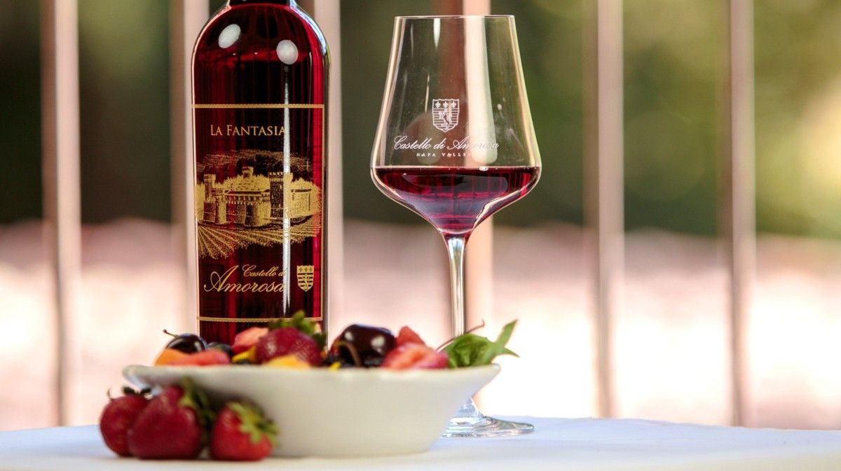 Bottle of red wine and glass with bowl of fruit on a table outside.
