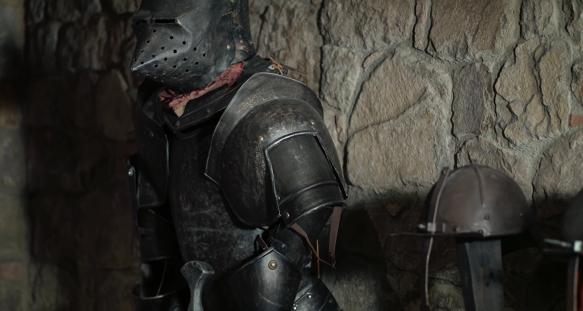 Knight in dark metal armor in a stone room, removing his helmet.