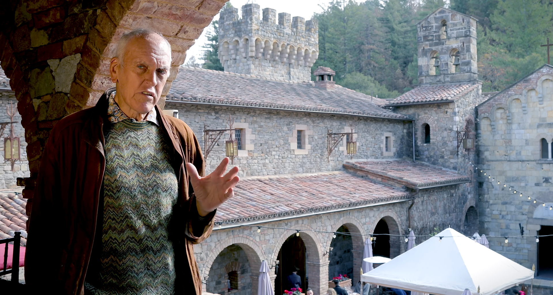 Man gesturing in front of a stone building with turrets and arched entrance.