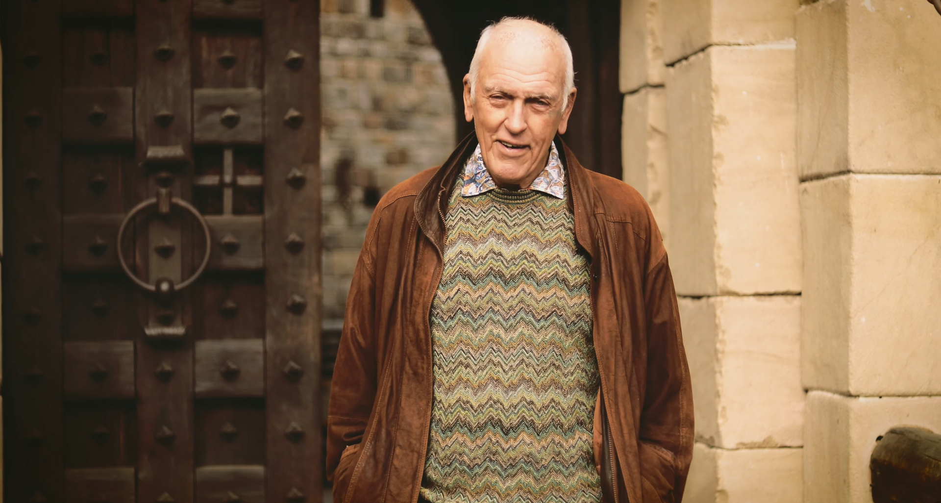 Older man, wearing brown jacket and sweater, stands in front of a large wooden door.
