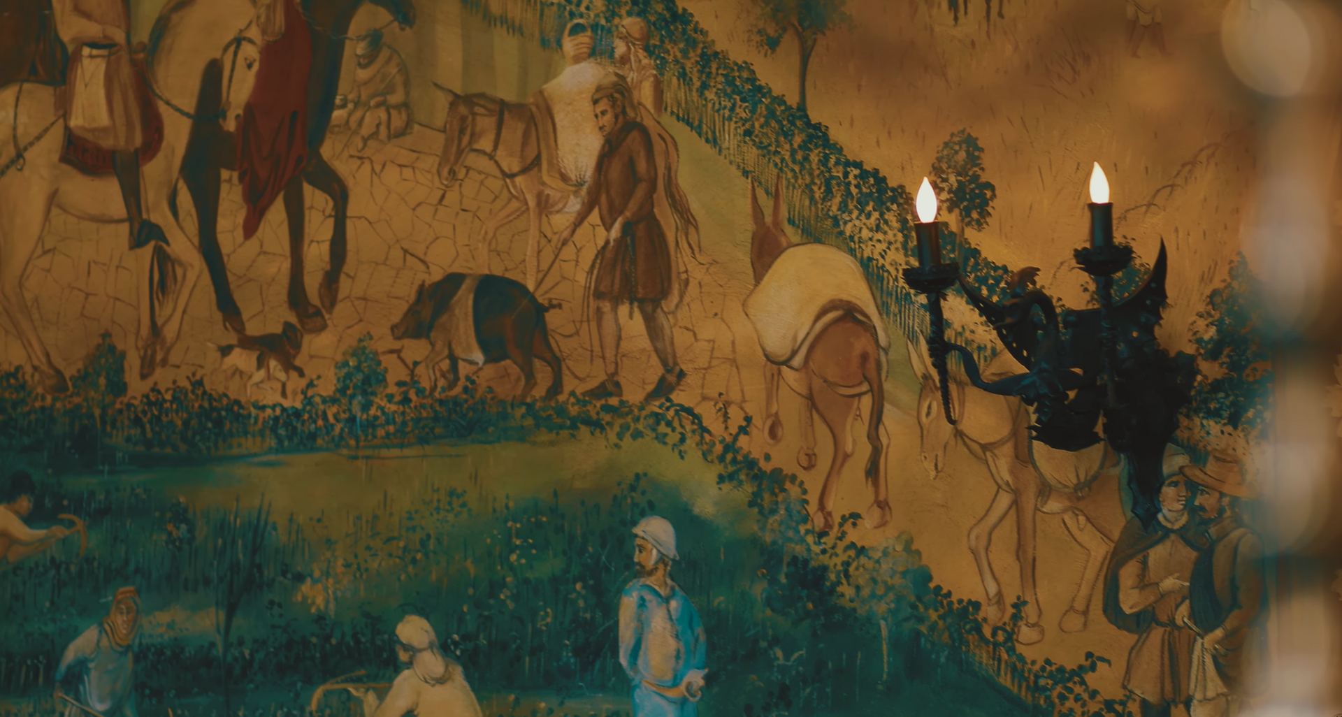 Tapestry depicting figures in historical clothing, with horses and a pig, lit by a sconce with flickering candles.