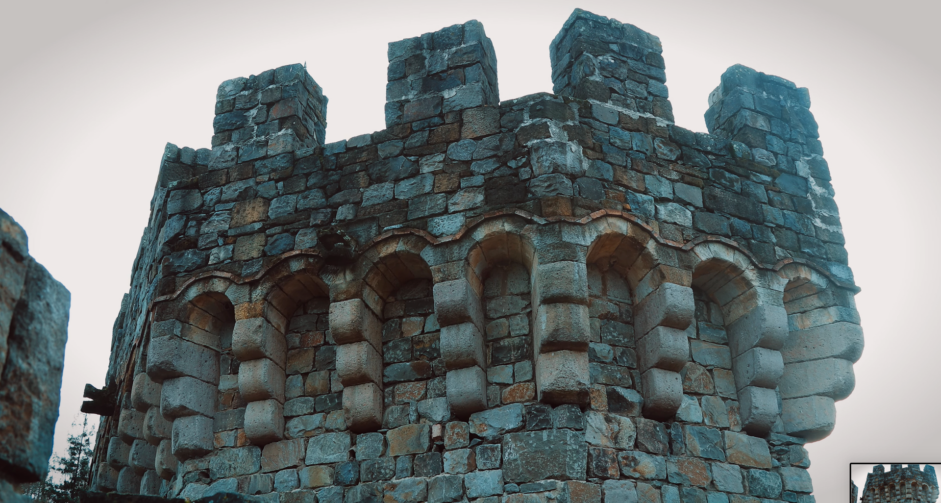 Stone castle tower with arched openings and crenellations against a cloudy sky.