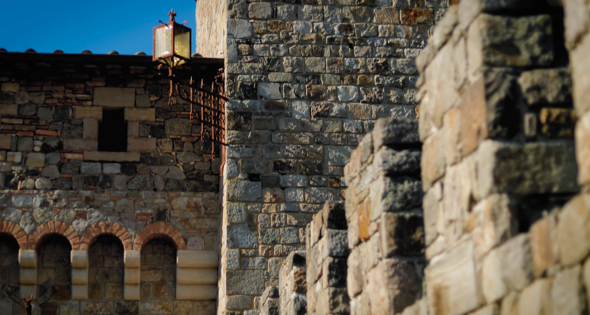 Stone walls of a medieval castle, with arched windows and a tower against a blue sky.