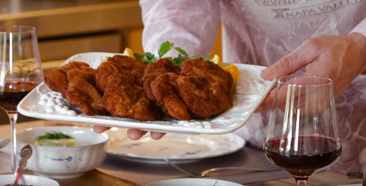Person presenting a platter of breaded cutlets, garnished with lemon and parsley, on a table set with wine and dishware.