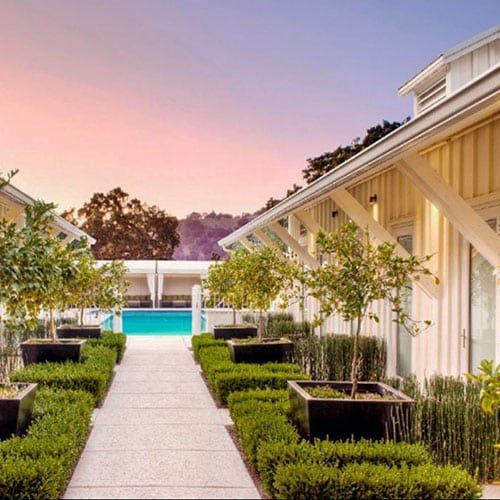 Pathway to a pool with trimmed hedges and trees, flanked by white buildings under a sunset.