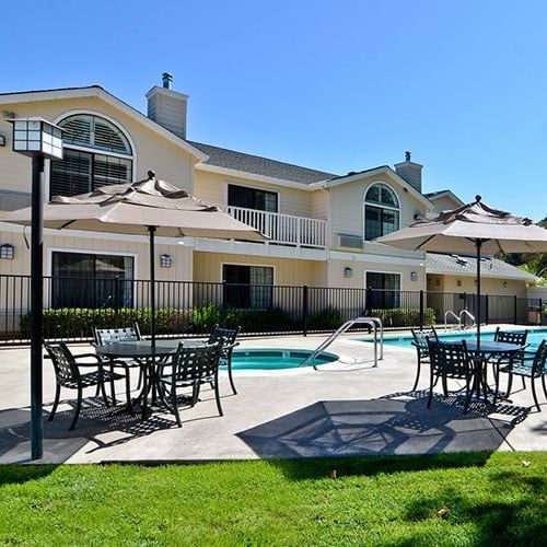 Poolside seating with umbrellas, pool, and beige building with black railings under a blue sky.