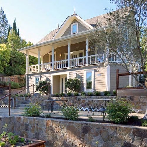 Two-story house with a wrap-around porch, tan siding, and stone retaining wall with outdoor seating.