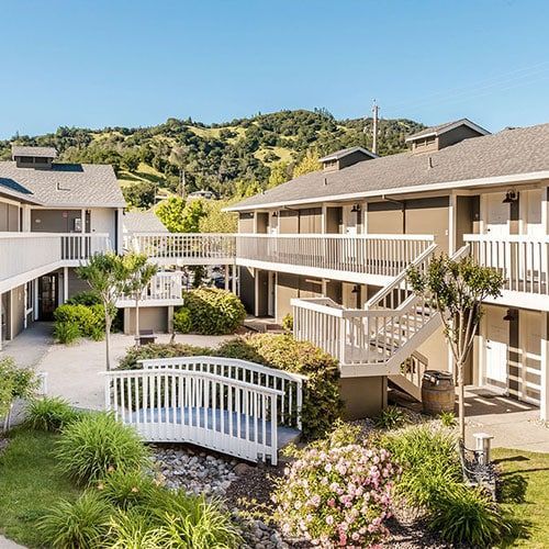 Courtyard with multi-story buildings, white railings, stairs, bridge, greenery, and hills in the background.