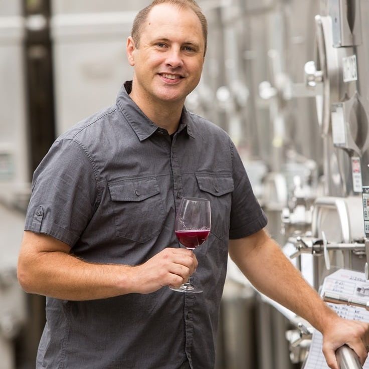 Man in gray shirt holds a glass of red wine, smiling near wine tanks.